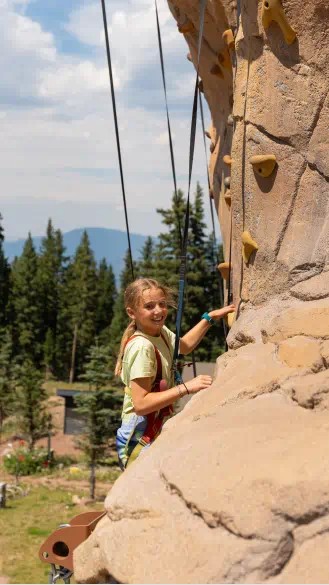 Little girl looks over an edge of rocks while she climbs on the outdoor climbing wall at camp aspen snowmass